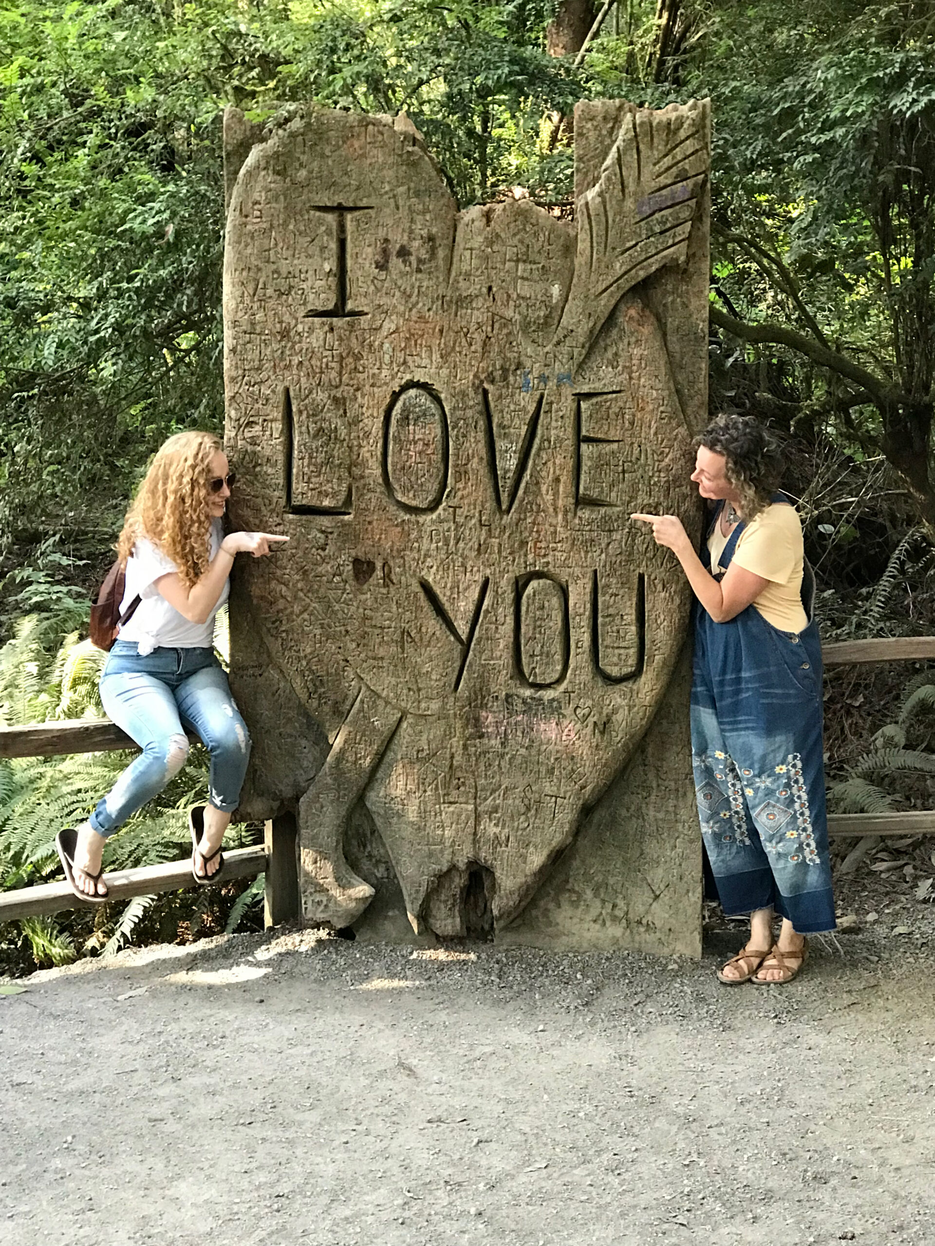 two women pointing to each other on either side of a large wood heart statue that says I Love you