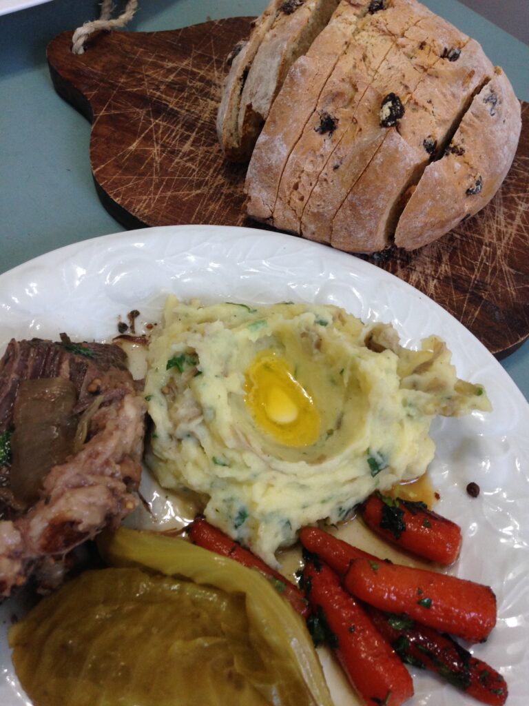 corned beef, cabbage, mashed potatoes, and carrots on a white plate with fig and fennel bread on a bread board