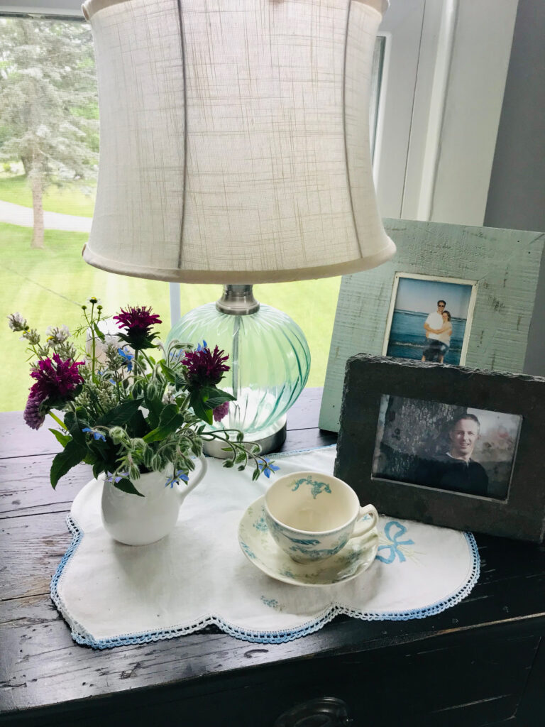 night stand with lamp, tea cup, photo frames and white pitcher filled with flowers