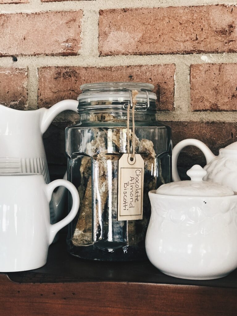 jar of chocolate almond biscotti on a mantle with white pitchers in front of a brick wall.