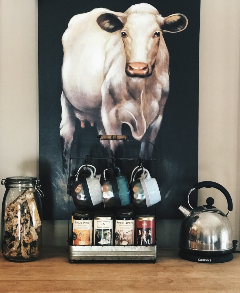 a mug tree with jar of chocolate almond biscotti and a tea pot with a cow picture behind it.