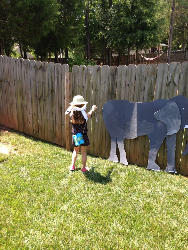 a girl pinning the tail on a cardboard elephant nailed to a fence