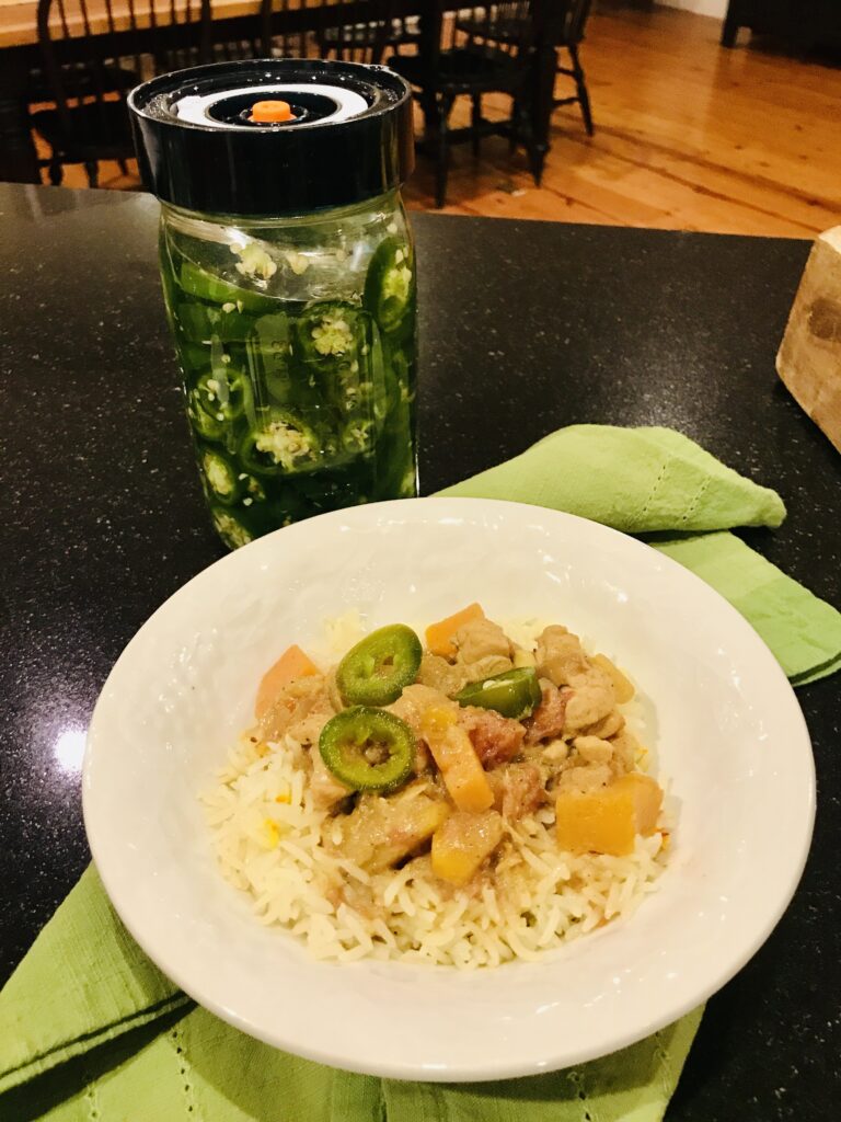 a bowl of chicken curry with fermented jalapeños, a jar of jalapeños and a green cloth napkin on a black countertop