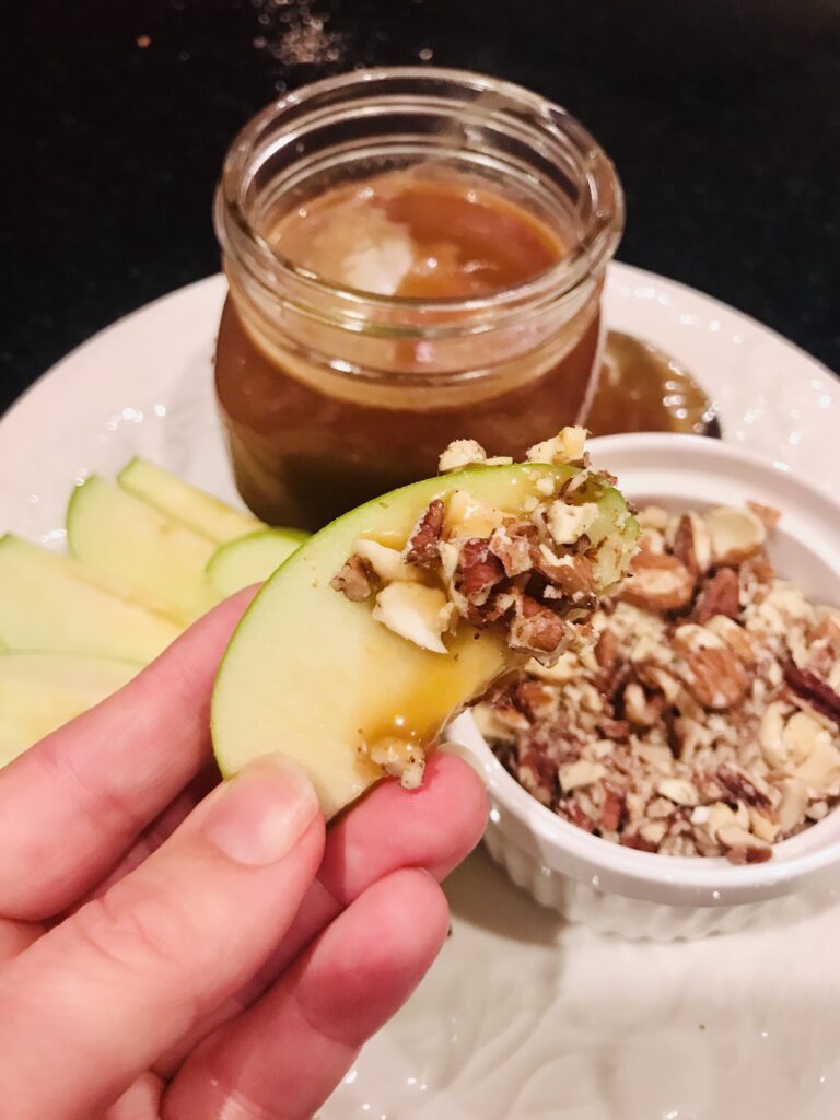 a hand holding an apple on a plate of apples, caramel sauce, and nuts