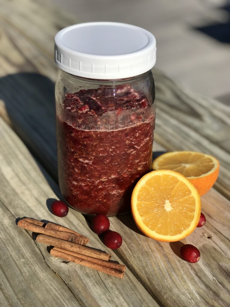 chutney in a jar on a wood table with orange, cinnamon stix, and cranberries