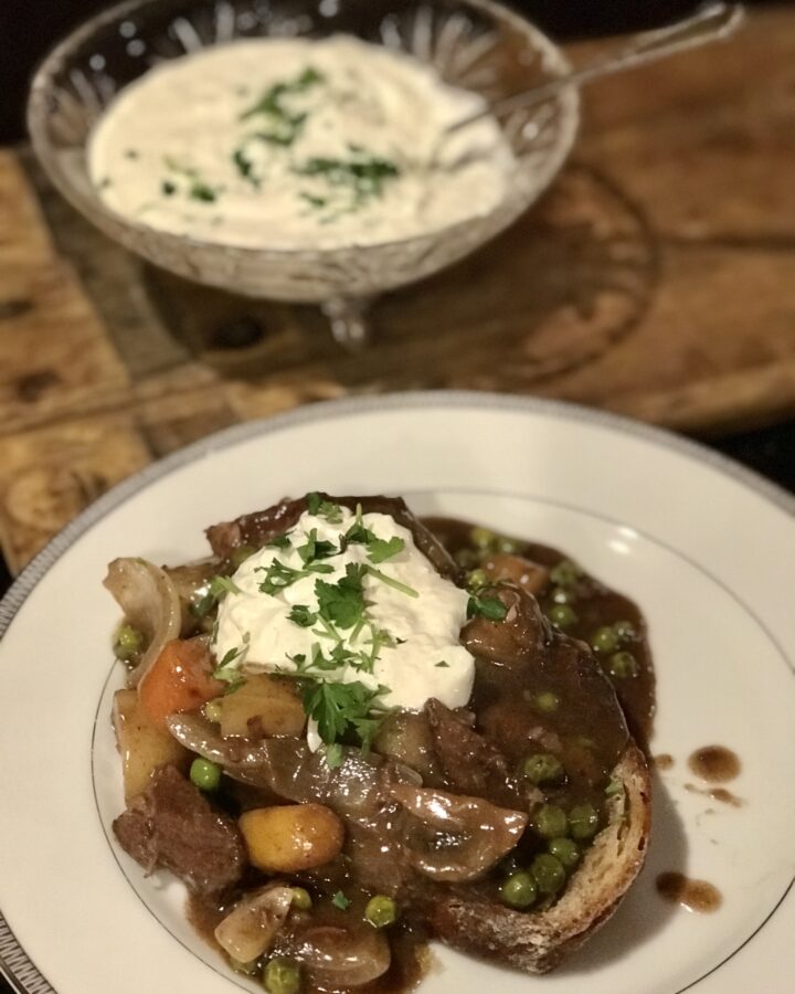plate of beef stew on peasant bread with cream sauce in the background