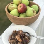 a bowl of apple crisp with a wooden bowl of apples behind
