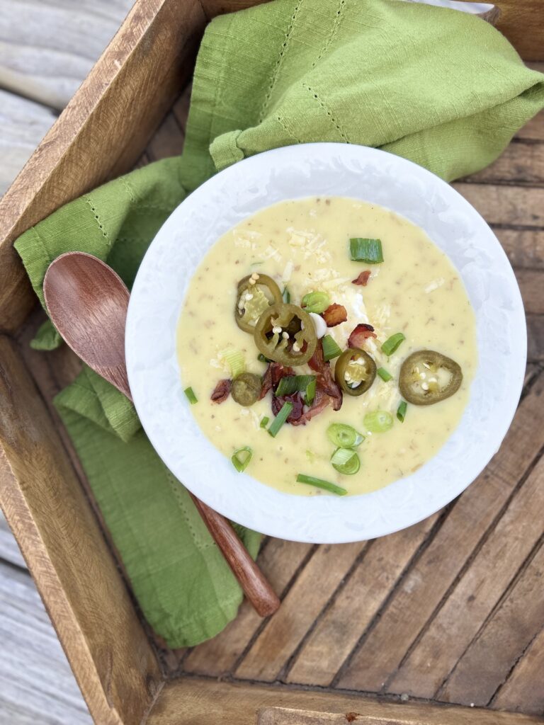 a white bowl of loaded baked potato soup on a wooden tray with a green napkin and a wooden spoon 