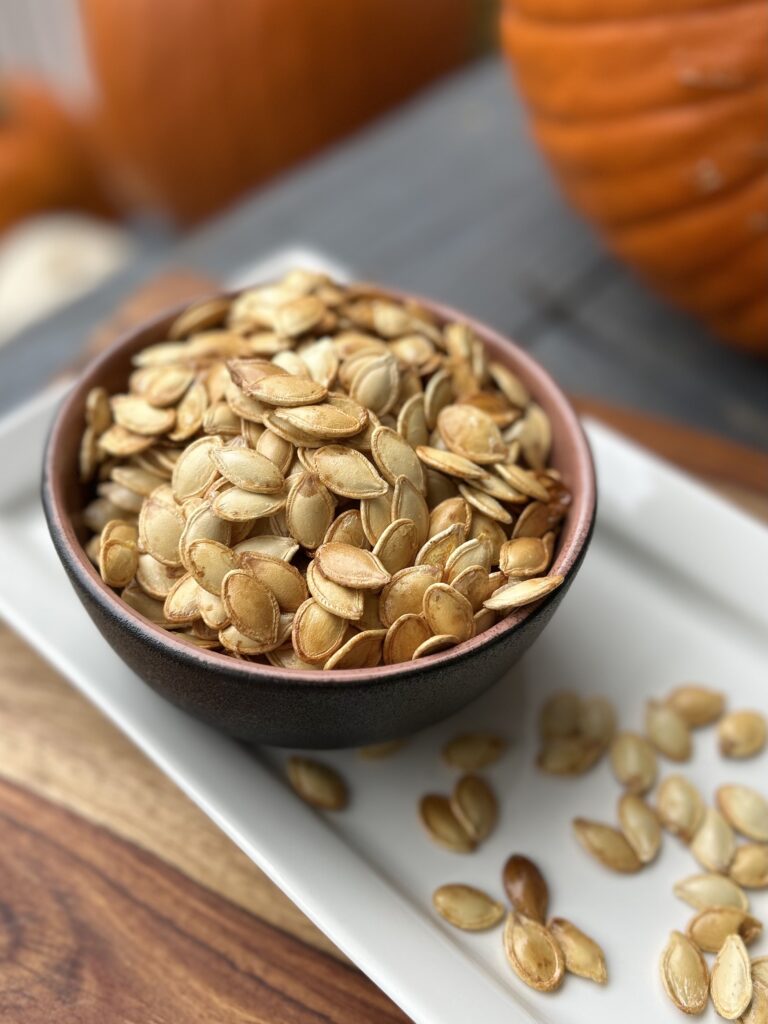 a bowl of roasted pumpkin seeds on a white plate with pumpkins in the background